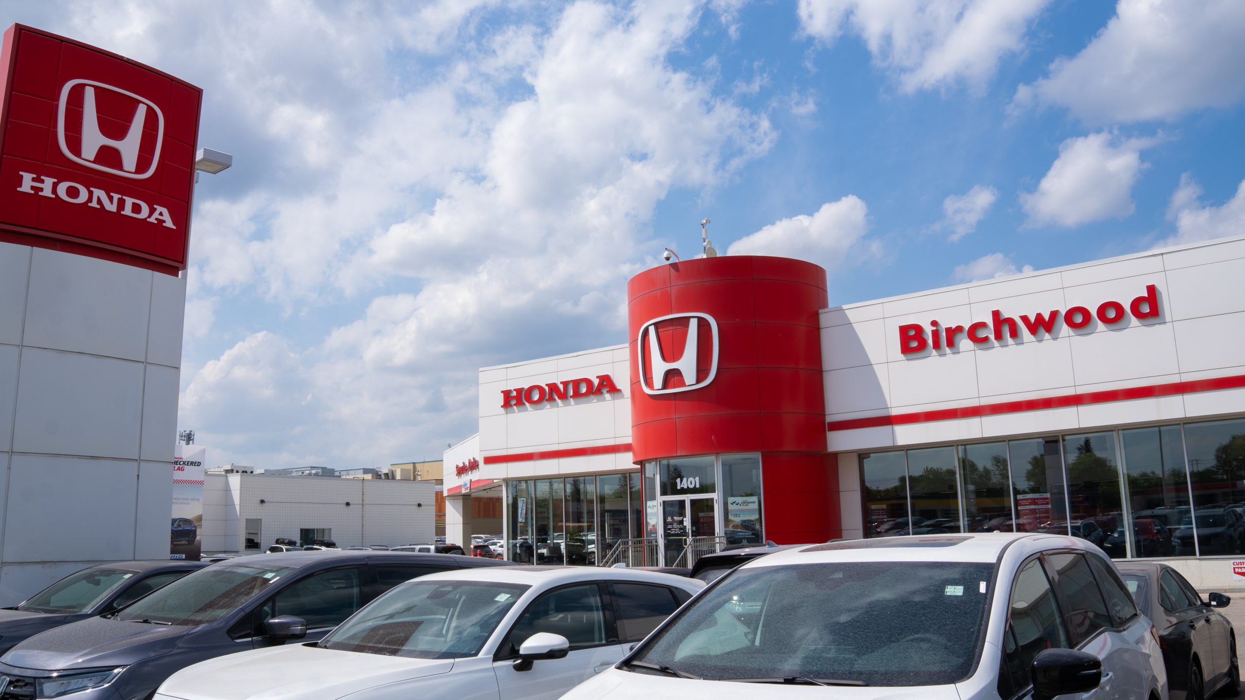 Front view of Birchwood Honda Centre featuring bold red Honda signage and branding, with several vehicles parked in the foreground under a partly cloudy sky.